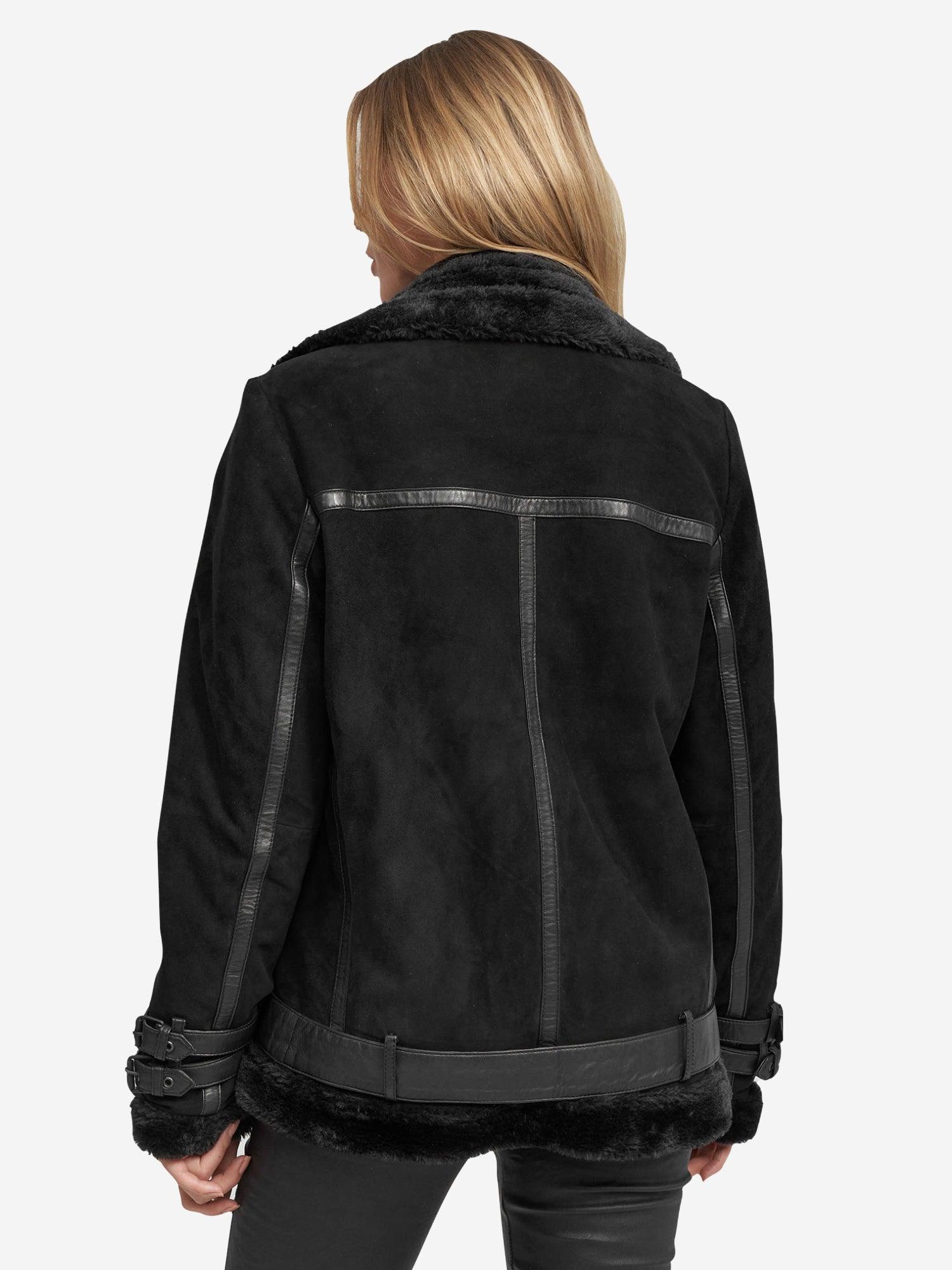 Von hinten ist eine blonde Person zu sehen, die eine trueprodigy Aloy Wilderlederjacke aus schwarzem Lammfell mit stylischen Schnallendetails an den Ärmeln trägt, ergänzt durch eine schwarze Hose, die sich vor einem schlichten weißen Hintergrund abhebt.