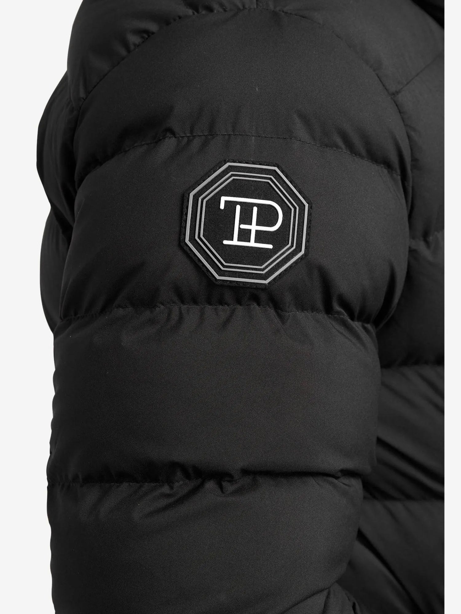 Die Chester Lange Pufferjacke mit Logo von trueprodigy zeigt eine Nahaufnahme eines schwarzen Ärmels mit einem sechseckigen Patch und einem weiß gestickten stilisierten Logo. Die gesteppte Textur unterstreicht ihre Qualität und sie verfügt über einen Reißverschluss für zusätzlichen Komfort.