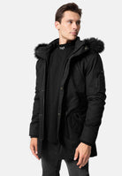 Mann trägt schwarze TRUEPRODIGY julius Winterjacke mit pelzbesetzter Kapuze und aufgesetzten Taschen; schwarzes Shirt mit „TPOY“-Aufschrift. Modernes Design, lässiger Stil, perfekt für kaltes Wetter.